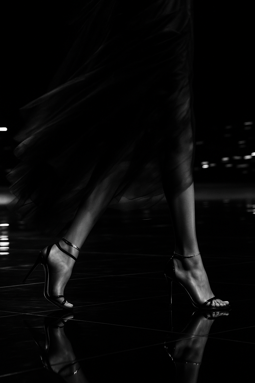 A close crop of heeled sandals and a flowing dress hem, caught mid-stride on a dark, reflective floor at night.